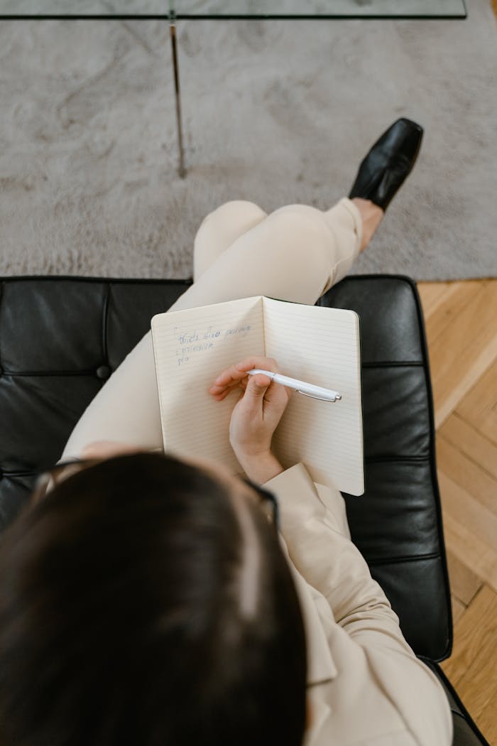 An overhead shot of a person writing in a notebook while seated on a black chair.